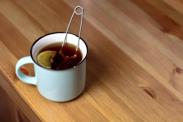 Hot warming tea with lemon in a light blue mug with a brewed tea strainer on a table with a wooden texture