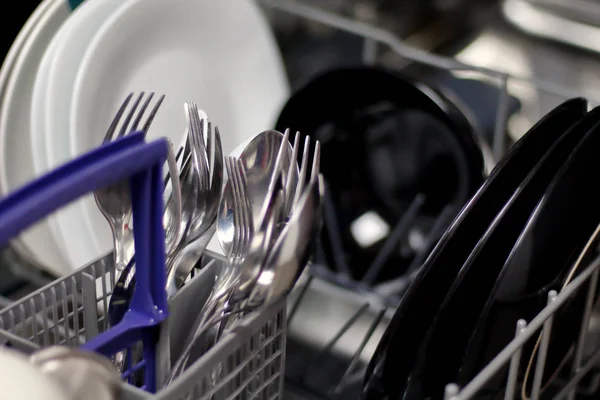 Dishwasher with white and black plates with bowls, forks, spoons, knives in a stand basket, clean washed dishes in a dishwasher