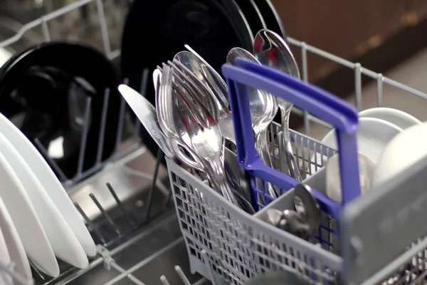 Dishwasher with white and black plates with bowls, forks, spoons, knives in a stand basket, clean washed dishes in a dishwasher top view
