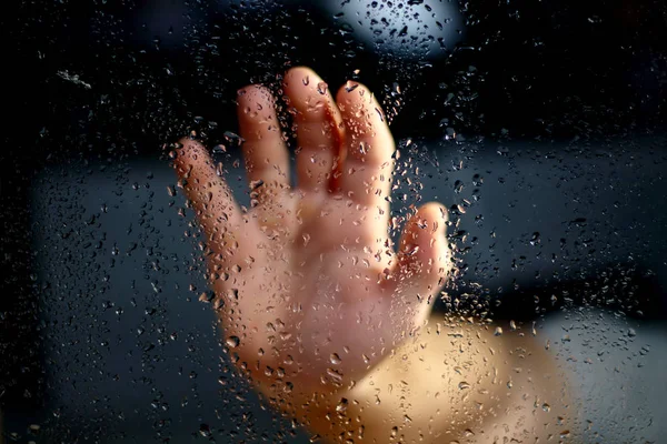 Male hand palm silhouette through the window with raindrops reaching for the glass. A request for help, depression blurred bokeh background