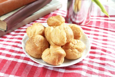 Kırmızı-beyaz kafes içinde rustik bir masa örtüsü ile bir masada çay ve kitap ile beyaz bir fincan yanında muhallebi ile ev yapımı küçük kek profiterole choux pasta
