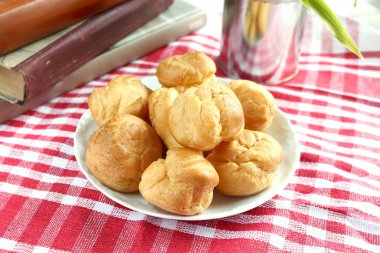 Kırmızı-beyaz kafes içinde rustik bir masa örtüsü ile bir masada çay ve kitap ile beyaz bir fincan yanında muhallebi ile ev yapımı küçük kek profiterole choux pasta