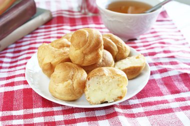 Kırmızı-beyaz kafes içinde rustik bir masa örtüsü ile bir masada çay ve kitap ile beyaz bir fincan yanında muhallebi ile ev yapımı küçük kek profiterole choux pasta