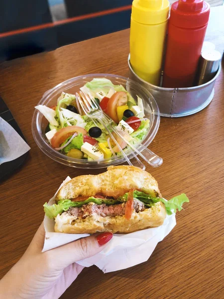 bitten hamburger in female hand in paper craft packaging in a fast food cafe with lettuce leaf, medium roast meat patty, tomato next to lettuce in a plastic container, unhealthy no diet food