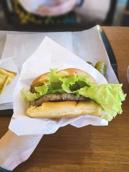 hamburger holding female hand in paper craft packaging in a fast food cafe with lettuce leaf, medium roast meat patty, tomato next to lettuce in a plastic container, unhealthy no diet food
