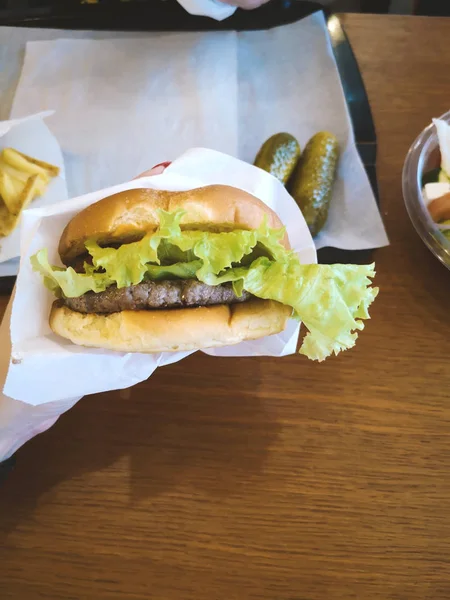 hamburger holding female hand in paper craft packaging in a fast food cafe with lettuce leaf, medium roast meat patty, tomato next to lettuce in a plastic container, unhealthy no diet food