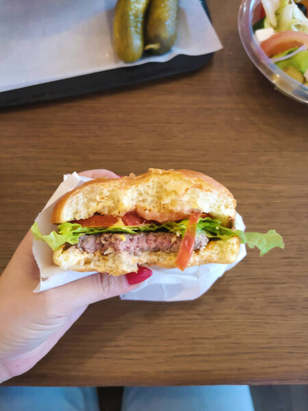 укушенный гамбургер в женской руке в бумажной упаковке ремесла в кафе быстрого питания с листьями салата, средняя жареная котлета, помидор рядом с салатом в пластиковом контейнере, нездоровая диетическая еда
