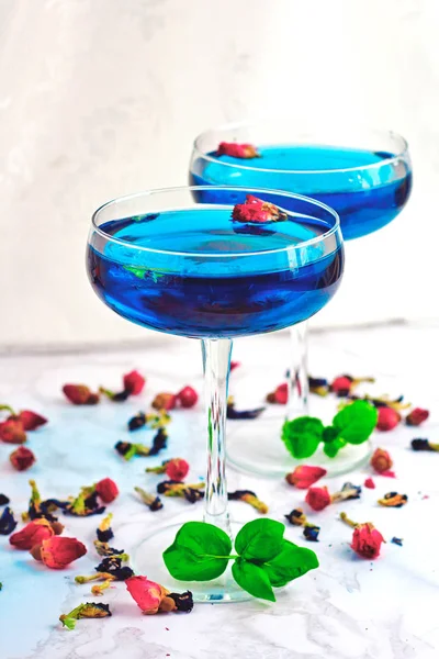 Thai asian blue Anchan tea in cocktail glasses with dry rosebuds and blue tea, tea leaves and green basil leaves, healthy organic detox Asian herbal tea clitoria Butterfly Pea on marble background