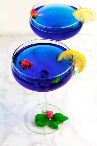 Thai asian blue Anchan tea in cocktail glasses with dry rosebuds, blue tea, tea and green basil leaves and slice of lemon, healthy organic detox Asian herbal tea clitoria Butterfly Pea on marble