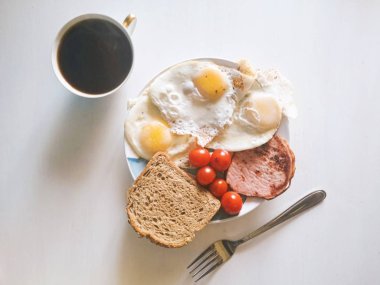 Sağlıklı organik dengeli kahvaltı, doğru beslenme, omlet, yumurta, domates kiraz, kepekli ekmek ve pastırma mavi-beyaz plaka üzerinde çatal ile yakın yatıyor, fincan siyah kahve üst görünümü. Instagram stili