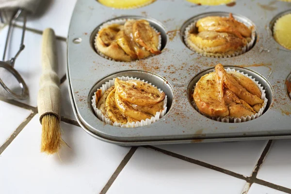 Gluten-free homemade vegetarian desserts cupcakes muffins with apple slices and pineapple rings on top in baking tray in kitchen on white-tiled worktop, next to a kitchen tea-strainer, towel