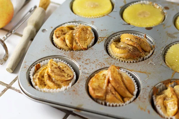 Gluten-free homemade vegetarian desserts cupcakes muffins with apple slices and pineapple rings on top in baking tray in kitchen on white-tiled worktop, next to a kitchen tea-strainer, towel