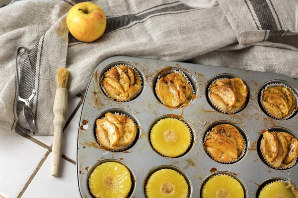 Gluten-free homemade vegetarian desserts cupcakes muffins with apple slices and pineapple rings on top in baking tray in kitchen on white-tiled worktop, next to a kitchen tea-strainer, towel