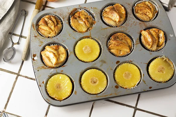 Gluten-free homemade vegetarian desserts cupcakes muffins with apple slices and pineapple rings on top in baking tray in kitchen on white-tiled worktop, next to a kitchen tea-strainer, towel