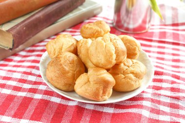 Isırılmış ev yapımı küçük kek karne choux pasta kırmızı-beyaz kafeste rustik bir masa örtüsü ile bir masada çay ve kitap ile beyaz bir fincan yanında muhallebi ile