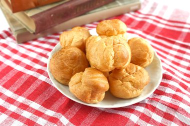 Isırılmış ev yapımı küçük kek karne choux pasta kırmızı-beyaz kafeste rustik bir masa örtüsü ile bir masada çay ve kitap ile beyaz bir fincan yanında muhallebi ile