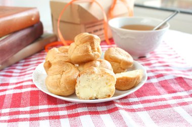 Isırılmış ev yapımı küçük kek karne choux pasta kırmızı-beyaz kafeste rustik bir masa örtüsü ile bir masada çay ve kitap ile beyaz bir fincan yanında muhallebi ile