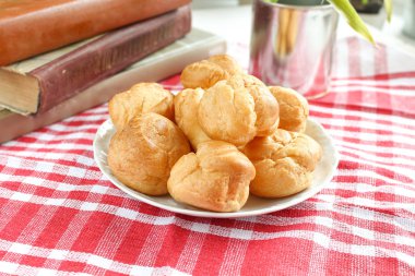 Isırılmış ev yapımı küçük kek karne choux pasta kırmızı-beyaz kafeste rustik bir masa örtüsü ile bir masada çay ve kitap ile beyaz bir fincan yanında muhallebi ile