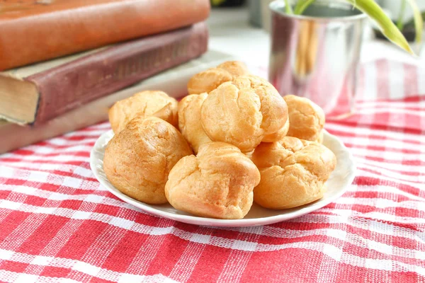 Isırılmış ev yapımı küçük kek karne choux pasta kırmızı-beyaz kafeste rustik bir masa örtüsü ile bir masada çay ve kitap ile beyaz bir fincan yanında muhallebi ile