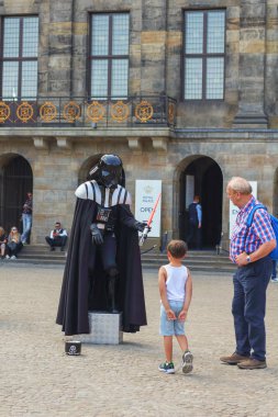 Amsterdam, Hollanda, Temmuz 2019: Darth Vader kostümlü kırmızı ışık kılıcı giyen aktör, Amsterdam'ın merkezindeki Baraj meydanında insanlarla ve çocuklarla konuşan ve oynayan heykelde yaşayan 