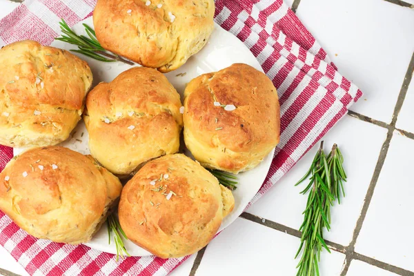homemade fresh herb bread rolls of Provencal herbs with salt and a sprig branch of rosemary. Bread rolls bakery style on white plate and red and white towel in a cage italian style