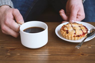 Gri kazaklı adam kışın kahve dükkanında siyah lungo americano kahvesi içer ve ahşap masada beyaz tabakta Amerikan ev yapımı vişneli turta yer. Sonbahar ruh hali instagram biçimi
