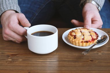 Gri kazaklı adam kışın kahve dükkanında siyah lungo americano kahvesi içer ve ahşap masada beyaz tabakta Amerikan ev yapımı vişneli turta yer. Sonbahar ruh hali instagram biçimi