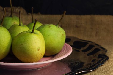 Yeşil organik armutlar ve diğerlerine benzemeyen küçük kırmızı bir elma gümüş metal tepside pembe bir tabakta, kaba retro bez kumaş brandasında pitoresk hayat. Karanlık atmosferik sonbahar hasadı