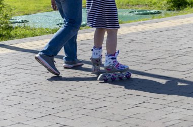 Bir adam kızının şehirdeki bir parkta paten lenmesine yardım eder. Geri görünüm