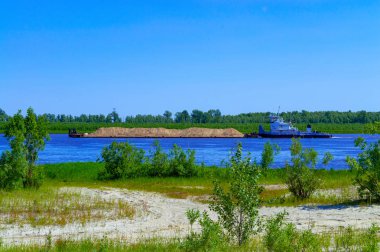 Bir römorkör Sibirya'da bir nehir üzerinde yüzer ve kum ile bir mavna iter. Yaz. Yeşil. Yan görünüm.