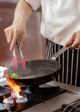 Aşçı, mutfak sobasında tavada alevle yemek pişiriyor.