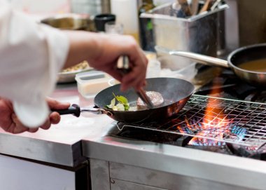 Aşçı, mutfak sobasında tavada alevle yemek pişiriyor.