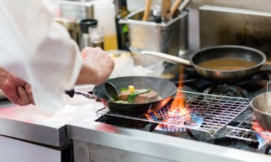 Aşçı, mutfak sobasında tavada alevle yemek pişiriyor.