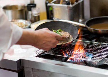 Aşçı, mutfak sobasında tavada alevle yemek pişiriyor.