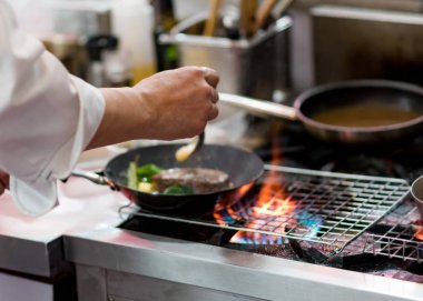 Aşçı, mutfak sobasında tavada alevle yemek pişiriyor.