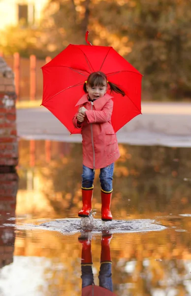 cute girl with tails in red rubber boots, trench coat with an autumn umbrella jumps in a deep puddle and smiles. Sunny autumn day. vertical photo of a funny child.