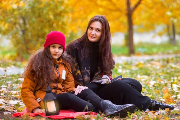Beautiful mother and daughter teenager on a picnic in autumn. A child in a red hat and an orange jacket, a woman in black clothes. Sitting under a tree on a plaid. In the background are yellow maple