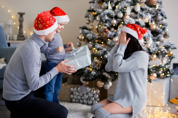 beautiful dad and son give mom a Christmas present, mom closed her eyes and a surprise awaits. family in gray clothes and red festive caps