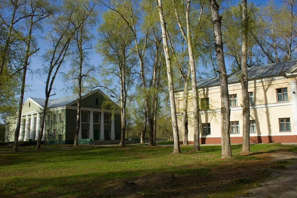 a large abandoned building of light yellow color in the Empire style stands on a green meadow. tall deciduous trees grow around.