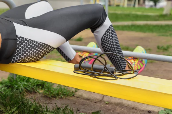 cropped shot of sporty woman in leggings with jumping rope sitting on bench outdoors and doing crunches