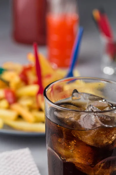 fizzy drink with ice in a glass on the background of a plate with fried potatoes. Close up.