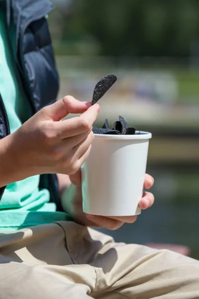 Black chips. Black potato chips in the hands of a teenager on the background of a sunny summer landscape.