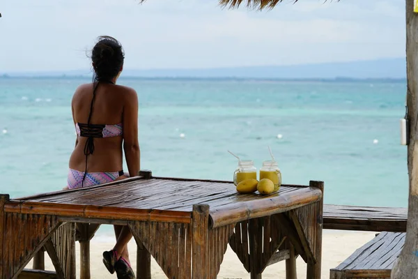 妇女坐在桌边与鸡尾酒在海滩上的背面视图 — 图库照片