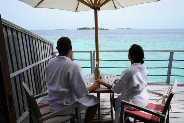 couple in bath robes sitting by table with sea view