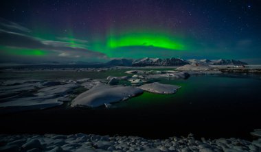 A beautiful green aurora borealis is dancing over the Jokulsarlon lagoon in the foreground is its reflection on the lakes surface. In the background are the northern lights above the mountains