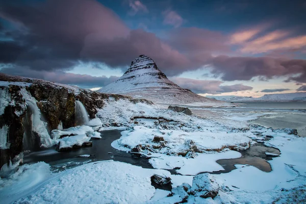 The sunrise at snowy Kirkjufell hill and frozen waterfall with morning aurora after long cold night with the northern lights. Winter arctic landscape. Iceland is country of snow and ice.