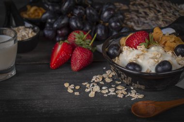 Yoğurt karışımı yulaf ezmesi, çilek ve ahşap masa üzerinde siyah kase tepesi kaşık, üzüm, çilek, süt cam, Mısır gevreği ve çevresinde yer yulaf ezmesi ile üzüm.
