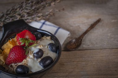 Yoğurt karışımı yulaf ezmesi, yanında yer kaşık ile kumaş ve ahşap masa mavi ve beyaz siyah kase çilek ve üzüm tepesi çizgili.