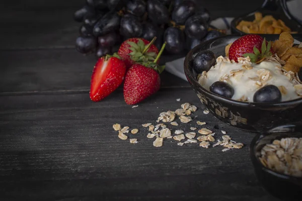 酸奶混合燕麦片 草莓和葡萄在黑色碗中的木桌与葡萄 玉米片和燕麦片周围放置 — 图库照片