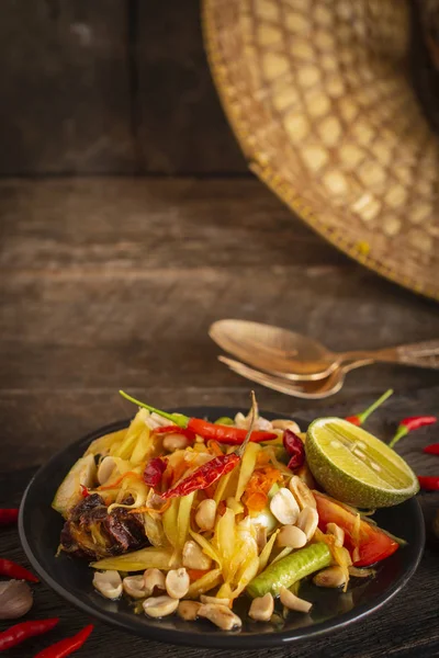 Салат папайя (Som tum Thai) на черной тарелке на деревянном столе расположены чеснок, вилка, ложка и чили . — стоковое фото